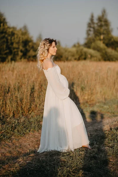 A young expectant mother in the third trimester of pregnancy in a white dress hugs her stomach against the backdrop of a natural landscape. The concept of future motherhood