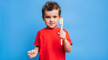 A smiling boy with healthy teeth holds a toothbrush on a blue isolated background. Oral hygiene. A place for your text