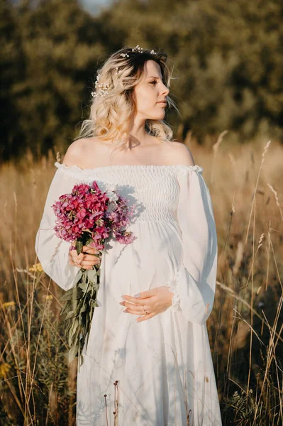 白いドレスに花の花束を手に妊娠3期の若い期待の母親は 自然の風景を背景に彼女の胃を抱擁します — ストック写真
