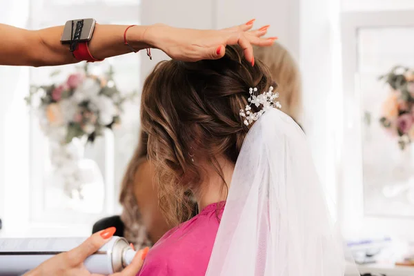 A hairdresser makes an elegant hairstyle for styling a bride with a white veil in her hair in the salon.