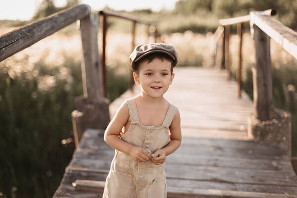 A boy is playing on a farm or ranch field, resting. Portrait of a child boy in a cap and overalls standing on the bridge. Childhood