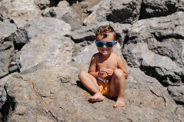 Dziecko Okularach Słonecznych Siedzi Opala Się Dużych Głazach Plaży Pobliżu — Zdjęcie stockowe