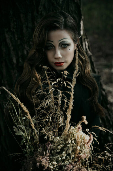 Portrait of a young woman in the Gothic gloomy image of a witch in the forest with a bouquet. Halloween costume. Vertical photo. Blur