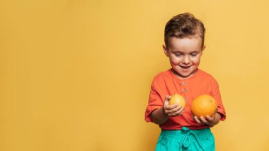 Sarı arka planda taze limon ve portakal tutan gülümseyen bir çocuğun stüdyo fotoğrafı. Sağlıklı bebek mamaları, vitaminler. Metnin için bir yer