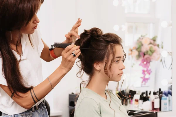 Shooting in a beauty salon. The hair master corrects the hairstyle of a young dark-haired girl with the help of a hair brush
