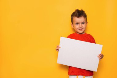Studio portrait of a happy boy with a clean white poster in his hands on a bright yellow background, with a place for your text or advertising.