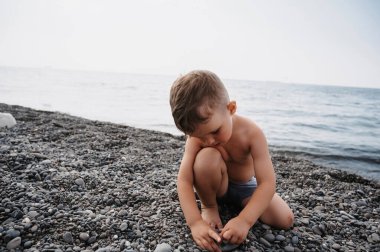 Tatlı bir çocuk kumsalda dinleniyor, deniz çakıllarıyla oynuyor. Deniz kıyısında açık hava aktiviteleri.
