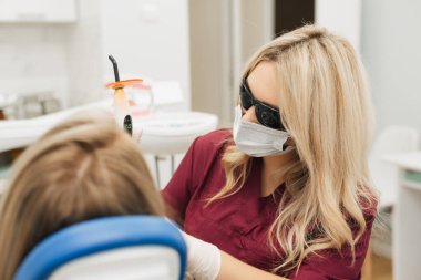 Profesyonel kadın doktor modern diş dolabındaki neşeli beyaz kadın için diş dolgusu yaptıktan sonra ultraviyole lamba kullanıyor..