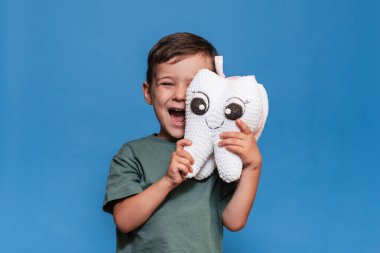 Sağlıklı dişleri olan gülümseyen bir çocuk mavi arka planda pelüş bir diş tutar. Ağız temizliği. Pediatrik dişçilik. Taşıyıcıların Önlenmesi