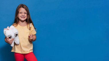 Sağlıklı dişleri olan gülümseyen bir kız mavi izole bir arka planda oyuncak diş ve diş fırçası tutuyor. Mesajın için bir yer. Ağız temizliği. Pediatrik dişçilik. Protezler. Diş fırçalama kuralları