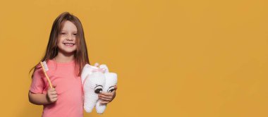 Sağlıklı dişleri olan gülümseyen bir kız sarı izole edilmiş bir arka planda oyuncak diş ve diş fırçası tutuyor. Mesajın için bir yer. Ağız temizliği. Pediatrik dişçilik. Protezler. Diş fırçalama kuralları