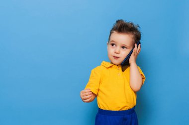 Küçük bir çocuk cep telefonuyla konuşuyor. Mavi arkaplanda stüdyo fotoğrafı.