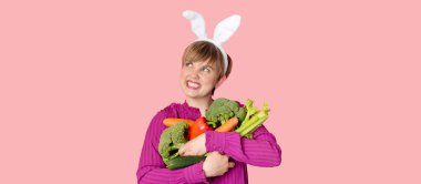 Stüdyo fotoğrafı, tavşan kulaklı beyaz bir kadın elinde bir sürü organik ve taze sebze tutuyor. Sağlıklı beslenme. Bir panoramik pankart ve bir uzay kopyası.