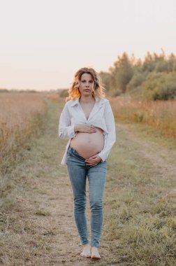 Bekleyen genç bir anne, batan güneşin ışınları altında doğal bir manzaranın arka planına doğru yürür. Hamilelik sırasındaki sakinlik kavramı. Üçüncü üç aylık dönem