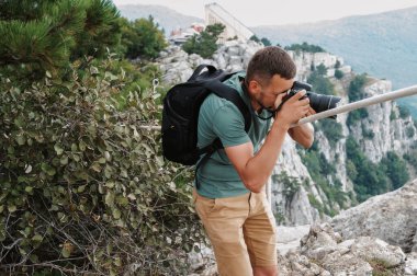 A young male tourist with a backpack climbed to the top of the mountain and takes pictures on the peak from a height. The concept of active life and hiking