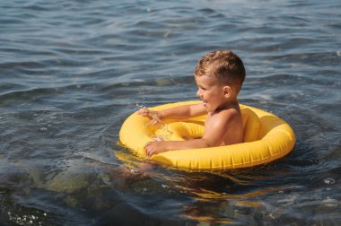 Sevimli erkek bebek, kumsalda denizde sarı şişme bir dairede yüzüyor.