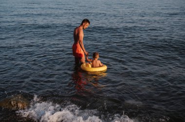 Baba çocuğa yüzmeyi öğretiyor. Deniz kenarında dinlenirken, baba ve çocuk denizde eğleniyor, yüzüyor ve suda oynuyorlar.