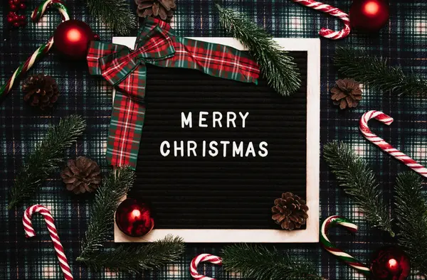 Eski bir mektup panosundaki plastik harflerle mutlu noeller. Arka planda karamel kamışlar, toplar, koniler ve köknar dalları var. Üst Manzara. Düz çizgi. Noel tebrik kartı.