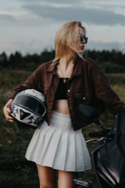 Motosikletin yanında bir kızın portresi, akşam, mavi gökyüzü, orman ve yolun arka planına karşı. Ellerinde bir kask tutuyor. Motosiklet yolculuğu