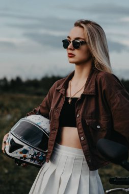 Motosikletin yanında bir kızın portresi, akşam, mavi gökyüzü, orman ve yolun arka planına karşı. Ellerinde bir kask tutuyor. Motosiklet yolculuğu