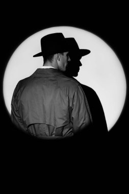 A dark silhouette of a male detective in a coat and hat in the noir style. A dramatic portrait in the style of detective films of the 1950s. A silhouette in a circle of light, like Agent 007