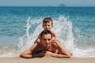 Mutlu bir baba ve oğul kumların üzerinde yatıyor. Çocuk babasının sırtüstü yatıyor ve dalgalar üzerlerini kaplıyor. Bir çocukla yaz günbatımı sahilinde tatil..