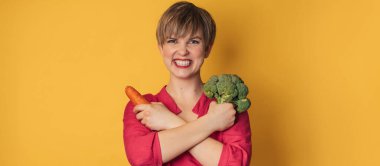 Sarı arka planda taze brokoli ve havuçla gülümseyen genç bir kadının stüdyo fotoğrafı. Yakın plan... Sağlıklı beslenme kavramı. Mesajın için bir yer. Panorama