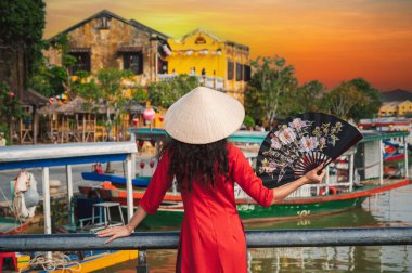 A woman in traditional Vietnamese cultural clothing in the ancient city of Hoi An on a bridge against the background of wooden boats decorated with lanterns clipart