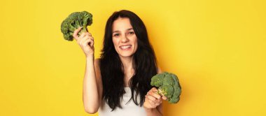 İnce, gülümseyen, beyaz, genç bir kadın sarı arka planda taze brokoli tutuyor. Panorama, metin için bir yer.