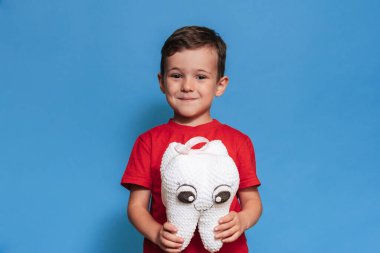 A smiling boy with healthy teeth holds a plush tooth in his hands on a blue isolated background. Oral hygiene. Pediatric dentistry. Prevention of caries. A place for your text