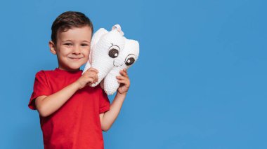 A smiling boy with healthy teeth holds a plush tooth in his hands on a blue isolated background. Oral hygiene. Pediatric dentistry. Prevention of caries. A place for your text