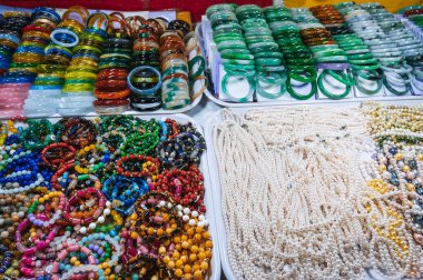 Bir pazarda sergilenen renkli, boncuklu bilezik ve kolye koleksiyonu, değerli taşlar, inciler ve gümüş elementler, geleneksel ve etnik mücevher tasarımları sergileniyor.