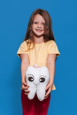 Sağlıklı dişleri olan gülümseyen bir kız mavi izole bir arka planda elinde bir diş oyuncağı tutuyor. Dikey fotoğraf. Ağız temizliği. Pediatrik dişçilik. Protezler. Diş fırçalama kuralları.