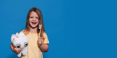 Sağlıklı dişleri olan gülümseyen bir kız mavi izole bir arka planda oyuncak diş ve diş fırçası tutuyor. Mesajın için bir yer. Ağız temizliği. Pediatrik dişçilik. Protezler. Diş fırçalama kuralları