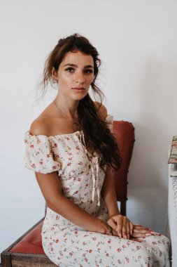 Klasik kadın elbisesi giymiş, eski mobilyaların üzerinde oturan genç bir kadının portresi.