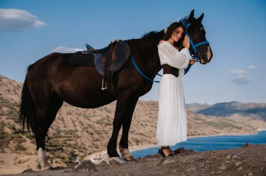 Beyaz elbiseli genç bir bayan bir dağ manzarasının arka planında atını nazikçe kucaklıyor. Romantizm konsepti, peri masalları