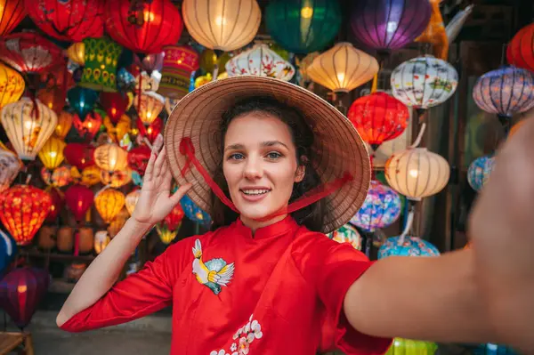 Antik Hoi An şehrinde fenerlerin arka planına karşı geleneksel Vietnam kıyafetleri ve konik Non La şapkalı bir kadının selfie 'si.
