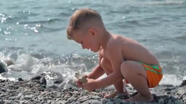 Deniz kıyısında taş ve çakıl taşlarıyla oynayan tatlı bir erkek bebek. Çocuk aile tatilinde iyi vakit geçiriyor. Yavaş çekim videosu.