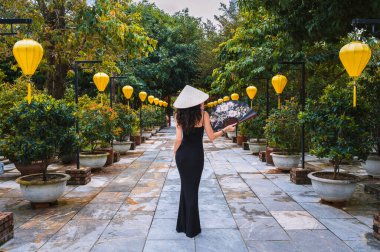 Geleneksel bir Vietnam şapkası takan ve arka planında fenerlerle süslenmiş Asya kültür bahçesinde yelpazesi olan bir kadın..