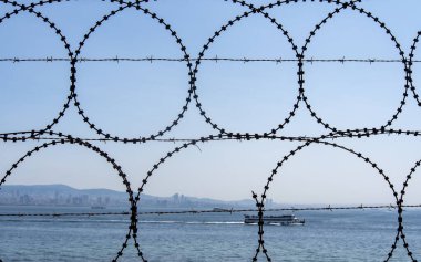 Sahil şeridi boyunca uzanan dikenli tel örgü, dingin suyun tam zıtlığını oluşturuyor. Bir tekne, açık mavi gökyüzünün altında pürüzsüz bir şekilde süzülür..
