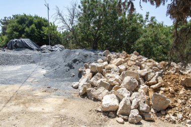 Ağır makineler, etrafı ağaçlarla çevrili bir şantiyeye malzeme taşıyor. Çakıl yığınları ve büyük kaya yığınları gün boyunca yoğun bir çalışma ortamını yansıtıyor..