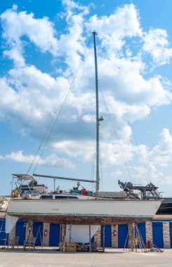 A sailboat rests on dry dock in a marina, surrounded by various vessels and bathed in bright sunlight with fluffy clouds scattered across the sky.