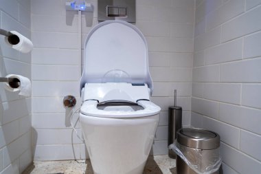 Şık, yüksek teknolojili bir tuvalet modern bir tuvaleti kaplıyor, bide seçeneği ve verimli bir tasarım sergiliyor. Yakınlarda, tuvalet kağıdı ve metal çöp tenekesi minimalist görünümü tamamlıyor..