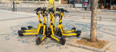 Sarı elektrikli scooter grupları güneşli bir günde kentsel hareketlilik seçeneklerini sergilemek için bir ağacın yakınındaki halka açık bir alanda düzgün bir şekilde park edilir. Yakınlardaki sokaklar canlı bir atmosfer olduğunu gösteriyor..