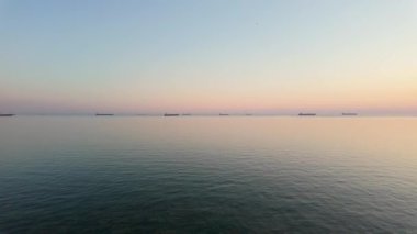 Sakin sular, şafağın yumuşak tonlarını yansıtır. Bazı gemiler uzakta demirlemiştir. Huzurlu atmosfer deniz kenarında yeni bir günün başlangıcını yakalıyor..