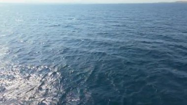 Güneş, derin mavi okyanus dalgalarının yüzeyinde parıldıyor. Gün ortasında huzurlu bir kıyı şeridinde sakin ve pitoresk bir atmosfer oluşturuyor..