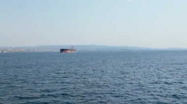 Uzak bir sahil kenti ufukta berrak bir gökyüzünün altında dururken büyük bir kargo gemisi sakin sularda yol alıyor. Huzurlu ortam deniz faaliyetlerini vurguluyor.