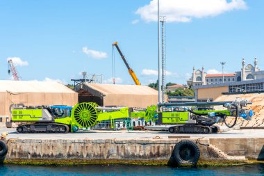 Yoğun bir limanın rıhtımında ağır inşaat makineleri kullanılmayı bekliyor. Parlak yeşil ekipman mavi sulara zıt düşüyor ve açık havada verimli işlemler sağlıyor.