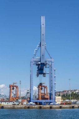 Rıhtımda belirgin bir mavi vinç dururken, daha küçük turuncu turnalar yoğun limandaki nakliye operasyonlarını destekliyor. Aktivite tüm hızıyla devam ederken gemiler yakınlarda demirledi..