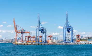 Vinçler, yoğun bir limanda çalışarak, bir rıhtım boyunca dik dururlar. Şehrin ufuk çizgisi açık mavi gökyüzünün altında yükseliyor..